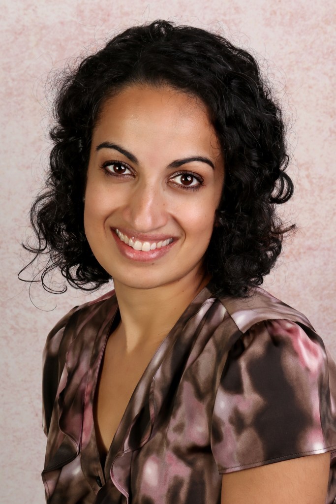 Dr. Aliya Chapman, a young brown woman with shoulder-length dark wavy hair. She wears a pink and brown patterned shirt in front of a textured beige background.