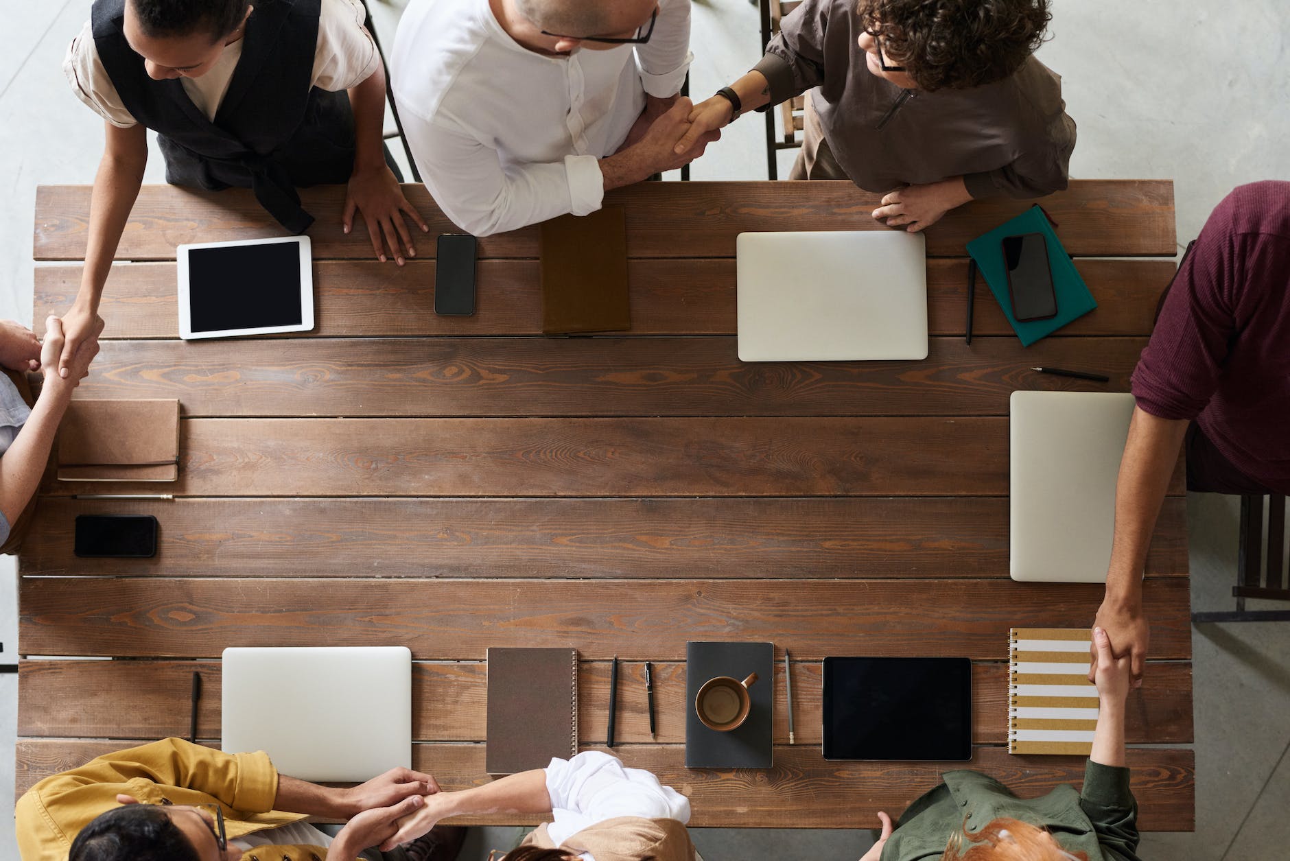 A group of people shake hands in pairs around a rectangular wooden table, notebooks and tablets laid out near them.