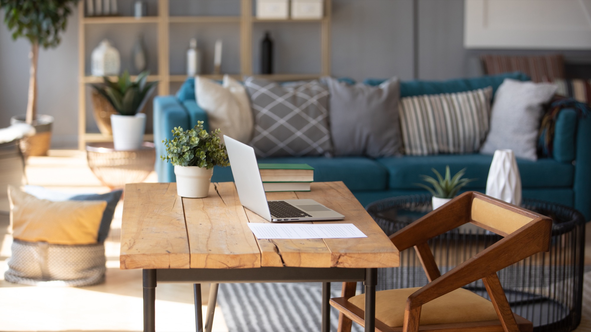 A laptop on a wooden desk in a living room with a teal couch and natural wood bookcase out of focus in the background.