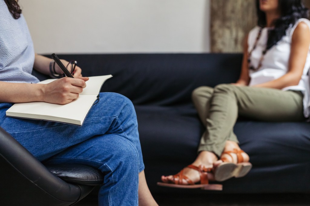 A woman wearing a grey shirt and jeans holds a pair of glasses and writes in a notebook. Nearby, another woman in a white shirt and olive green pants sits on a couch.