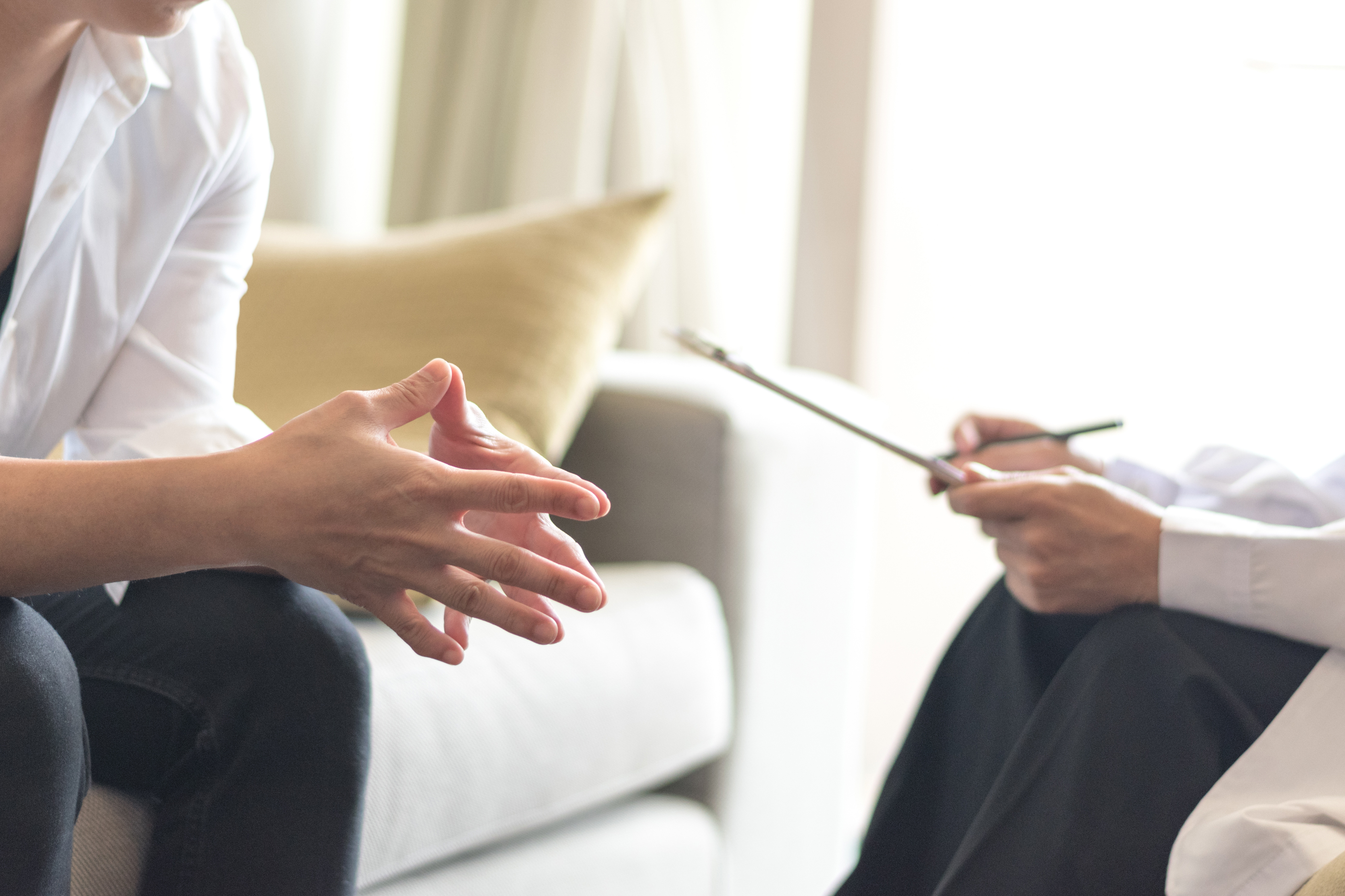 A person sitting on a couch leans forward, fingers steepled together, while another person in a chair nearby writes on a clipboard.